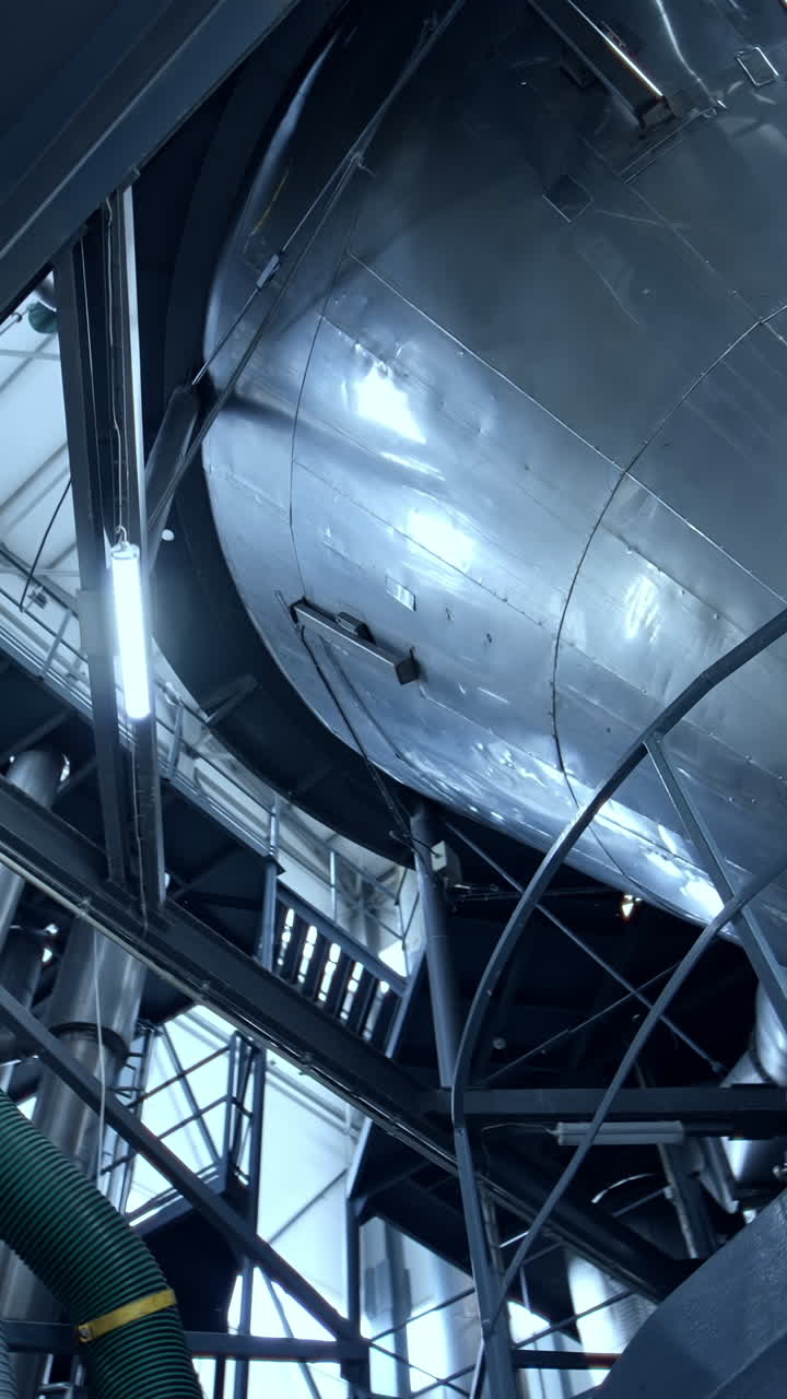 Huge dairy tank beside the thick plastic tubes. Stainless steel tank standing at the raised platform with sensors at its basics. Circle view from below. Vertical video