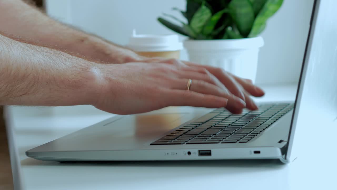 Male hands print the text on the laptop keyboard on a white table, a green indoor plant on the background. Without a face, a close -up
