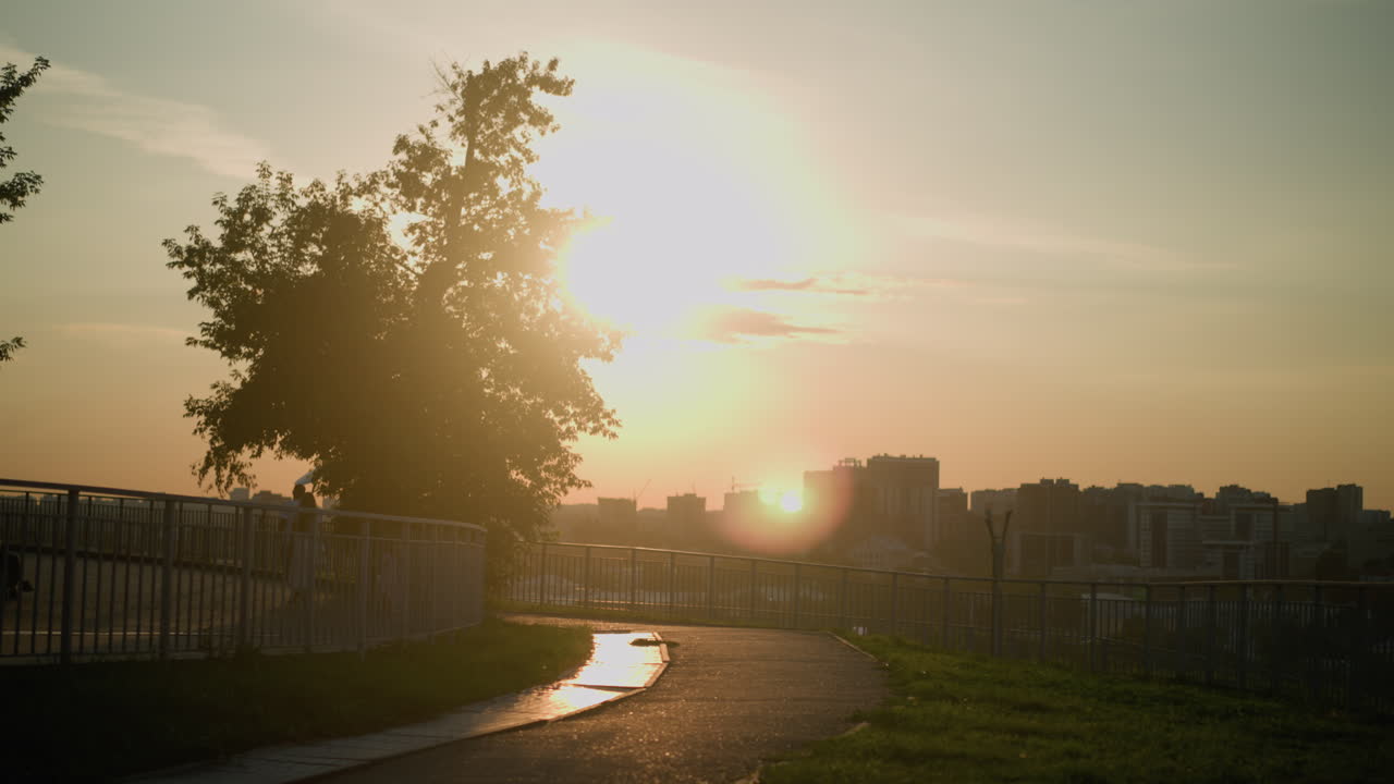 vista del paisaje urbano al atardecer con personas caminando por el camino cerca de la barandilla de hierro, grandes árboles proyectando sombras, la cálida luz del sol iluminando los edificios urbanos en el fondo
