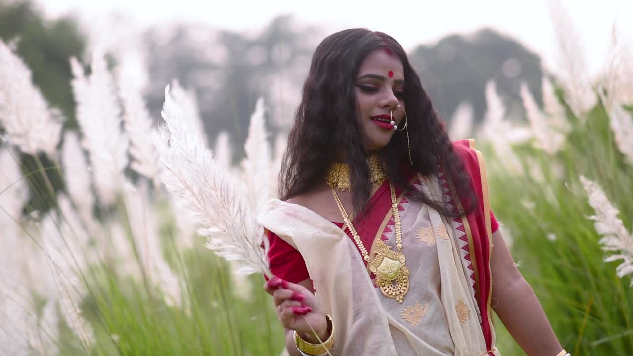 A cute and carefree newly wed Indian woman is happy and plays with white flowered grass in a field on a windy day at sunset or surise