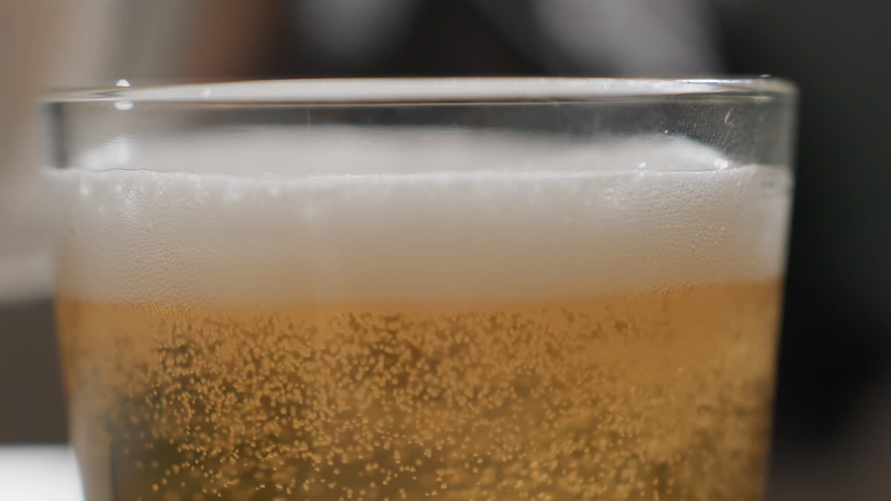 Primo piano di birra versata in un bicchiere con schiuma e testa che sale, liquido dorato che riempie il boccale, si forma uno spesso strato spumoso, bollicine vivaci e condensa, servizio del barista in un pub accogliente, tintinnio festoso.