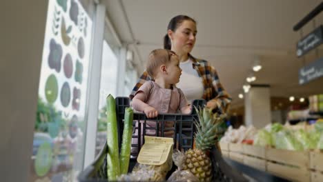 Interested-little-girl-baby-rides-in-a-cart-and-helps-her-mom-shopping-in-a-supermarket.-A-brunette-woman-and-her-little-daughter-choose-goods-during-their-shopping-in-a-supermarket