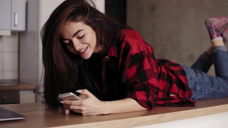 una atractiva chica morena está soñando y sonriendo sobre algo mientras está acostada en la superficie de la mesa de la cocina y enviando mensajes de texto a alguien.