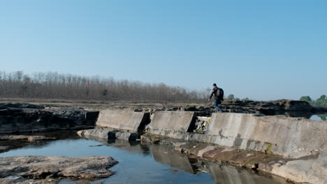 unrecognizable man with backpack crossing river over bridge rubble