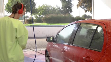 woman cleaning roof of red car with pressure hose while listening to music with mask on