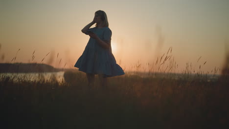 woman in a blue gown, swaying gently, enjoys a sunset while using a smartphone and wearing a headset in a glowing, sunlit field