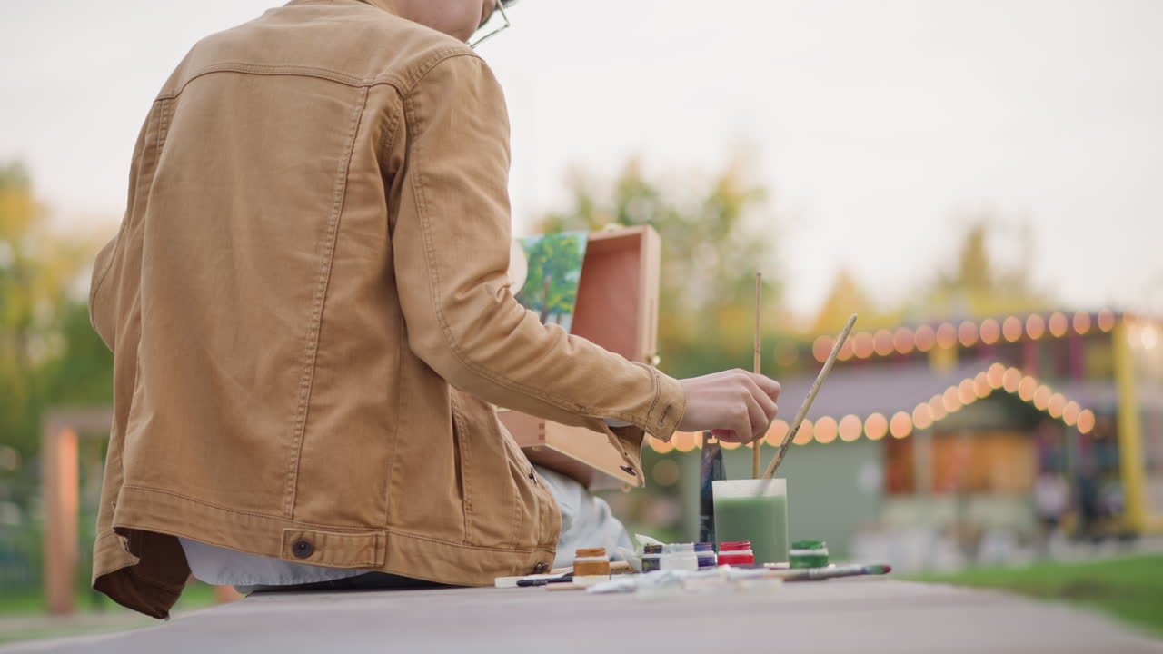 Artistic creation at park. Painter focused at outdoor festival table with glowing lights. Creative individual engaged in painting during relaxed outdoor festival scene with warm lighting