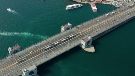 Public-Transport-Tram-Train-passing-Galata-Bridge-over-Bosphorus-in-Istanbul-with-Boats-on-water,-Scenic-Aerial-low-angle-follow-shot