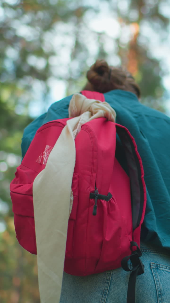 평화로운 빛의 숲을 고 있는 여자의 뒷모습, 어깨에 빨간 배을 걸고, 녹색 셔츠가 나무를 통해 빛이 필터링되는 자연과 여 있습니다.