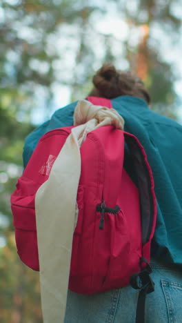vista trasera de una mujer caminando por un pacífico bosque iluminado por el sol, una mochila roja envuelta en su hombro, una camisa verde mezclándose con la naturaleza mientras la luz del sol se filtra a través de los árboles