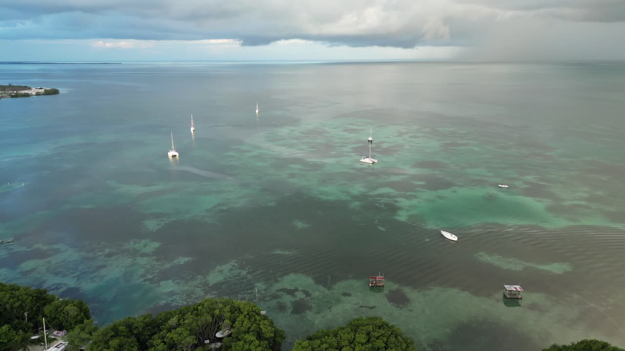 vista de avión no tripulado en belice volando sobre el mar azul oscuro y claro del caribe con barcos, un cay de arena blanca cubierto de palmeras en un día nublado