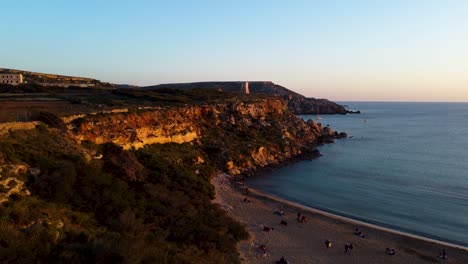 golden bay beach malta during golden hour sunset, sand rock cliff ridge, landmark tower on the hill, sea shoreline mediterranean hazy blue pink pastel orange glow horizon