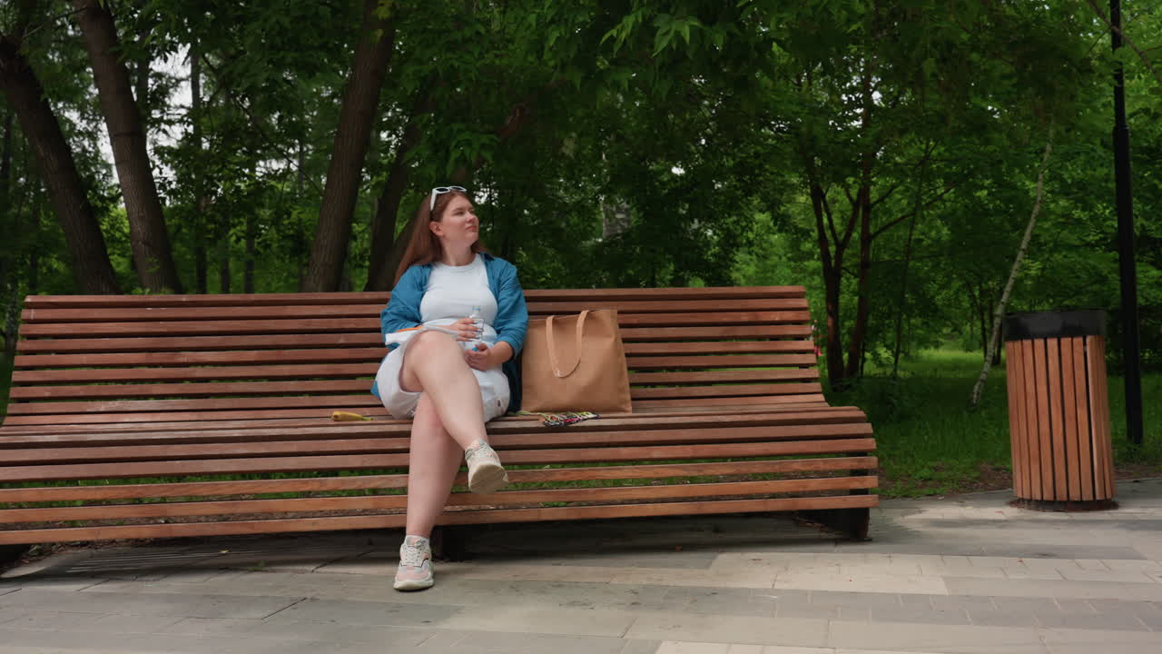 Student seated on wooden park bench with crossed legs gazes upward thoughtfully while chasing flies, relaxing in peaceful green park setting with bag beside her under soft daylight