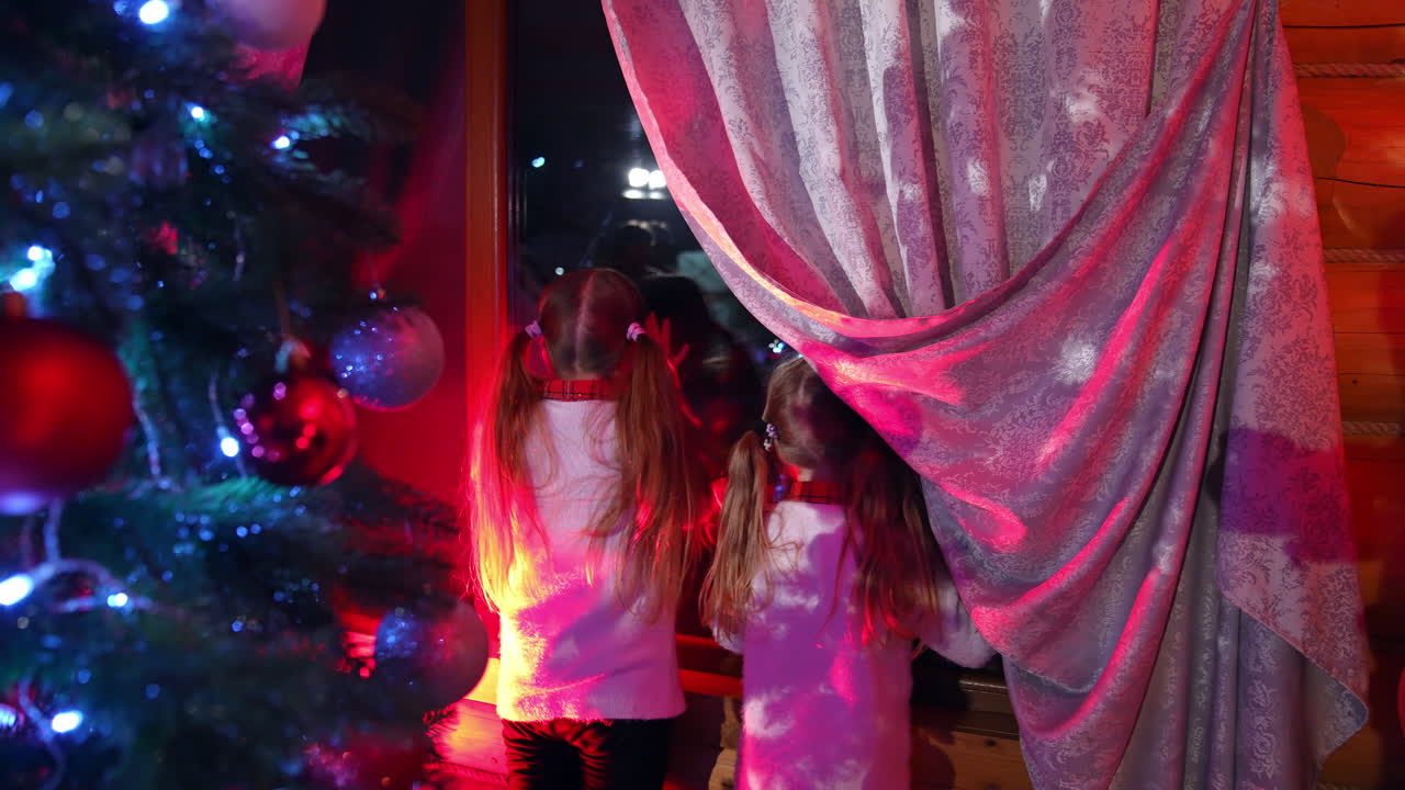 Kids waiting for Santa Claus at Christmas night. Two little girls looking through the window for Santa in the living room with beautiful Christmas tree