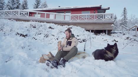 man and his pet dog sitting on snow next to pile of wood at winter