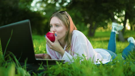 woman lying on grass using laptop while eating apple, deeply focused on work, leg raised casually in lush green surroundings with trees and bright natural light