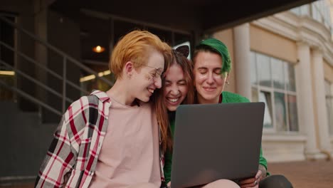 Happy-and-very-cheerful-girls-with-short-haircuts-of-bright-colors-in-checkered-shirts-and-their-friend,-a-brunette-girl,-are-looking-at-something-in-the-screen-of-a-gray-monitor-and-hugging-each-other