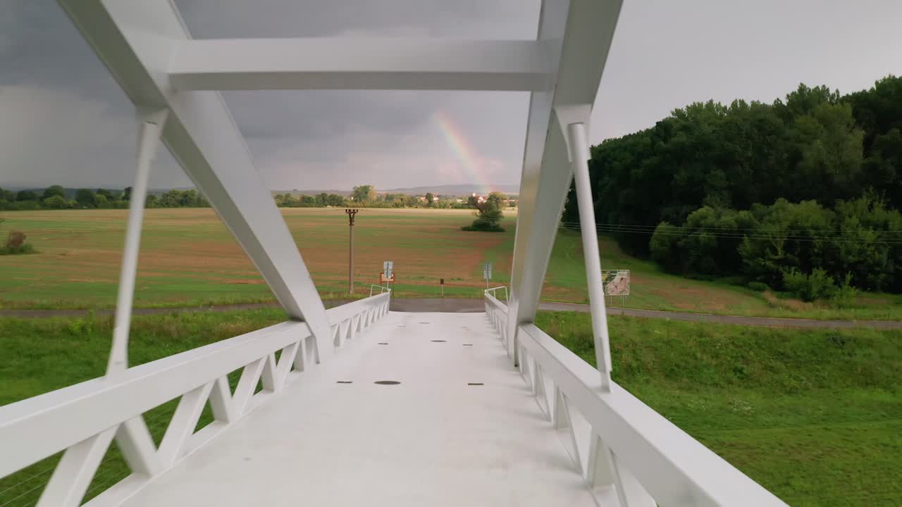 Flying fast forward through a cycling bridge running across calm river Moravia in Slovakia. Aerial sunset footage taken just before a storm with huge clouds and a rainbow in the background