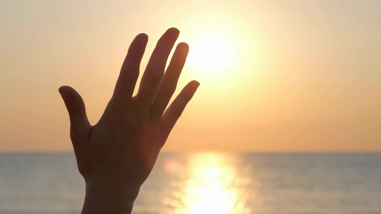 A single silhouetted hand rises in the foreground against a warm sunset over the sea, with shimmering reflections, a clear horizon, and a peaceful coastal