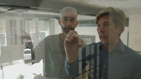 two mature male coworkers discussing scheme on glass wall in office