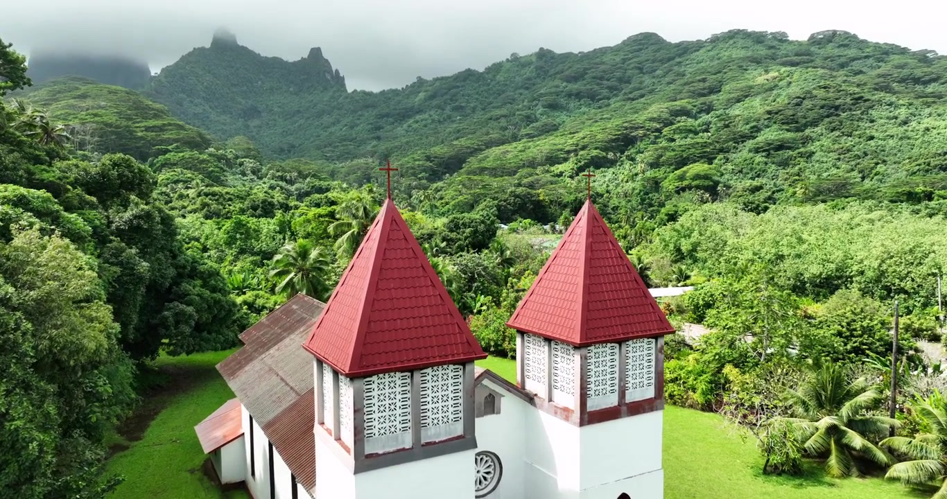 Campanarios gemelos de techo rojo se elevan desde la fachada de una iglesia misionera blanca en la Polinesia Francesa, una densa selva tropical verde rodea un césped cuidado bajo montañas escarpadas y brumosas. Toma de dron aérea de aproximación.