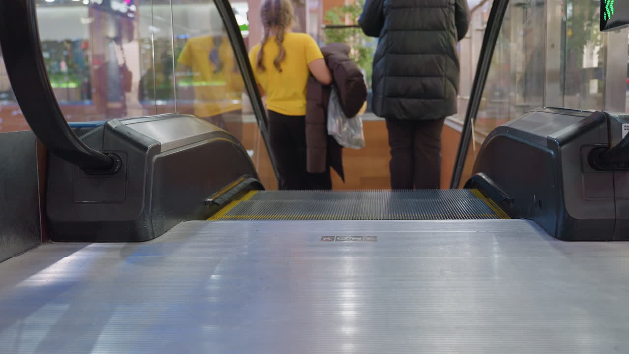 Back view of people in winter clothes stepping off descending escalator inside shopping mall with glass railing and bright lights reflecting modern indoor retail lifestyle