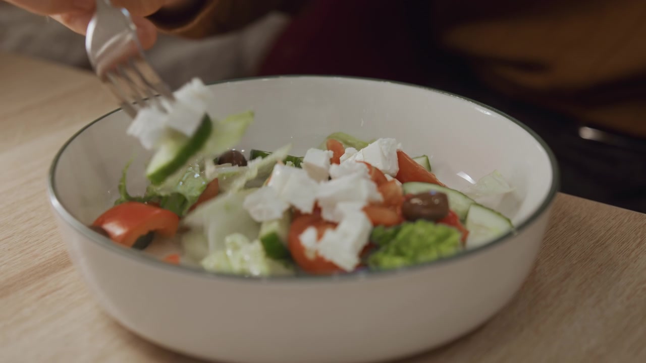 Close up of person eating salad with fork featuring fresh vegetables and feta cheese in white bowl on wooden table. Hand holding silver fork repeatedly picks food from plate while background