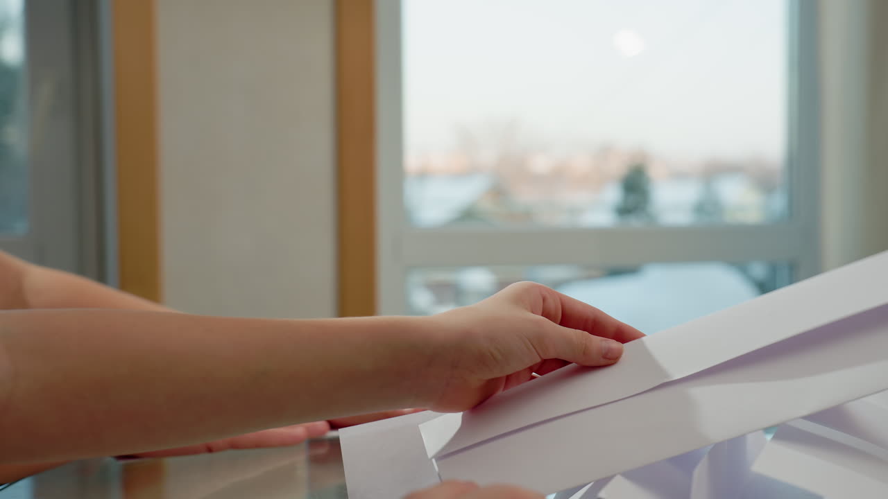 Hand view of kids folding white plain paper at table, background showing blurred window with snow outside, focus on hands and paper, creating a quiet and cozy crafting environment