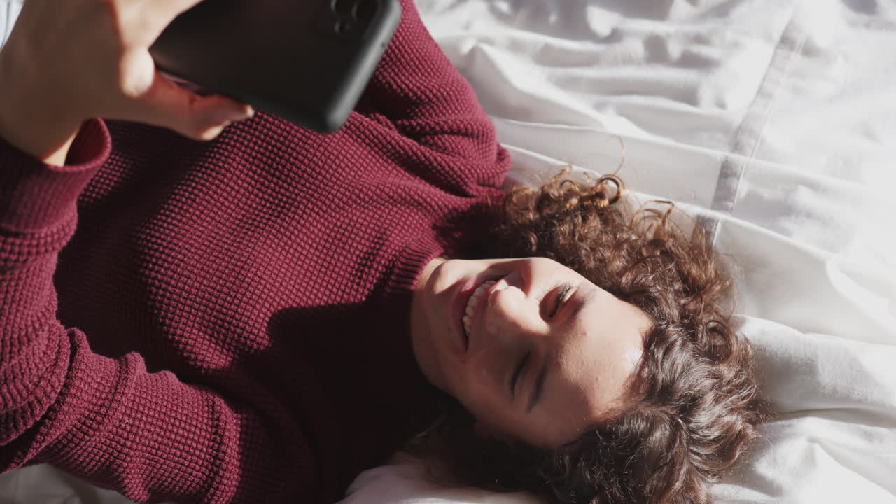 Slapper af i sengen, kvinde smiler mens hun bruger smartphone til underholdning.