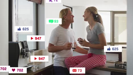 Animation-of-social-media-icons-and-numbers-flying-over-caucasian-couple-kissing-and-in-kitchen