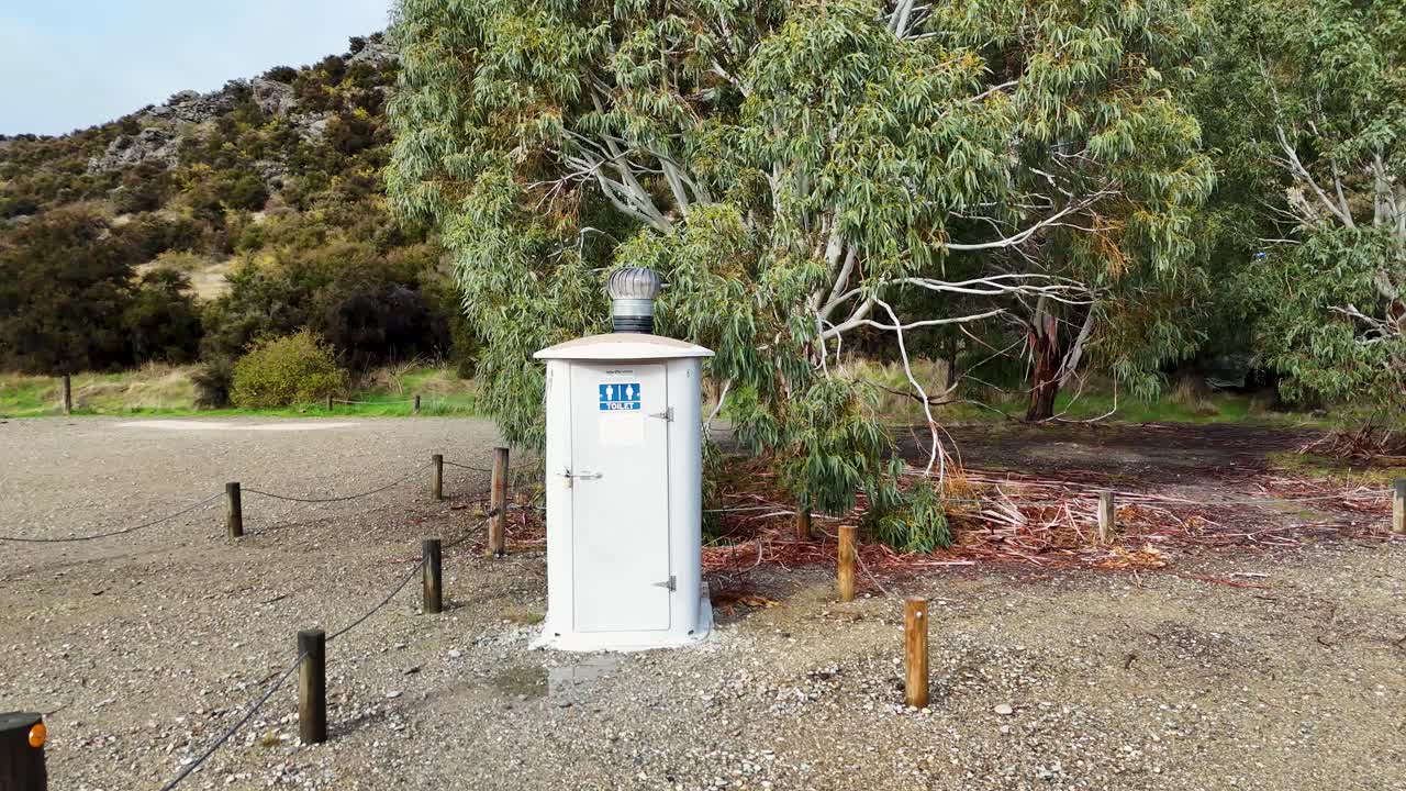 Drone camera smoothly pans across a solitary public toilet in a gravel parking area, surrounded by trees and hills under natural daylight