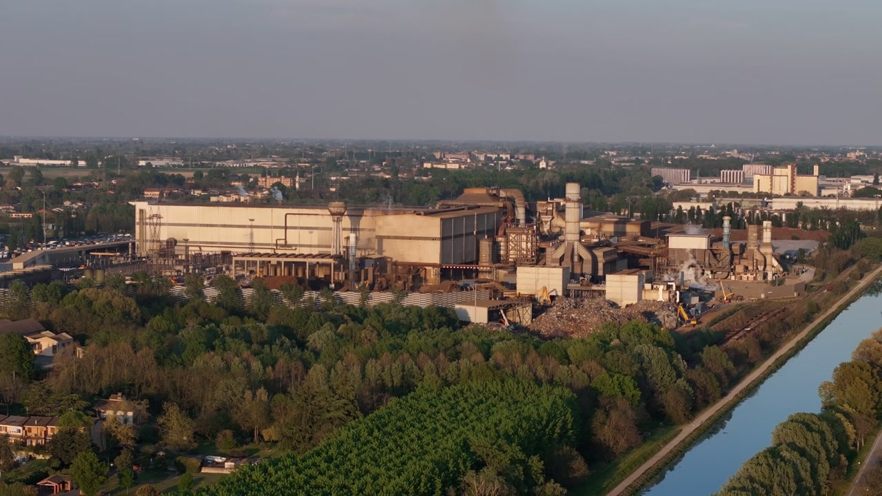 Acciaieria a Cremona, Lombardia, Italia, accanto al canale Idrovia Milano-Cremona, con acciaieria, strutture industriali e terreni agricoli della Pianura Padana, ripresa lenta in allontanamento con drone.
