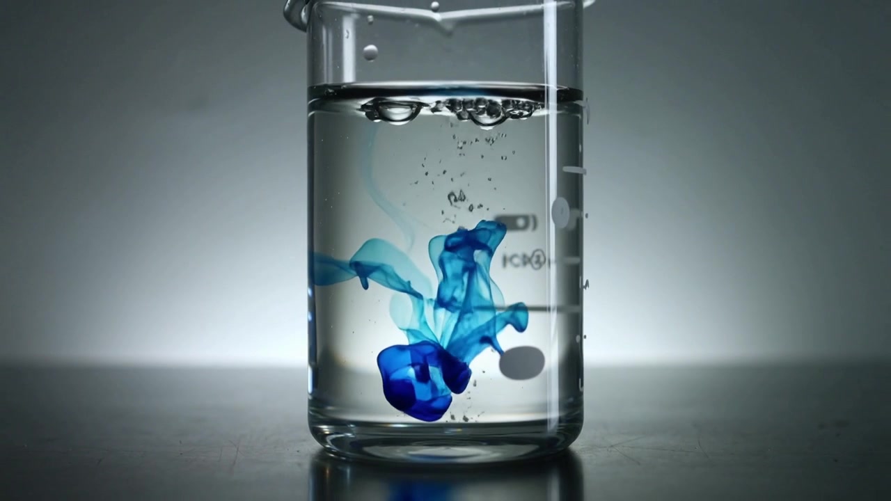 Goccia blu brillante che cade forando la superficie dell'acqua in un becher di vetro sul banco di laboratorio, mostrando la miscelazione