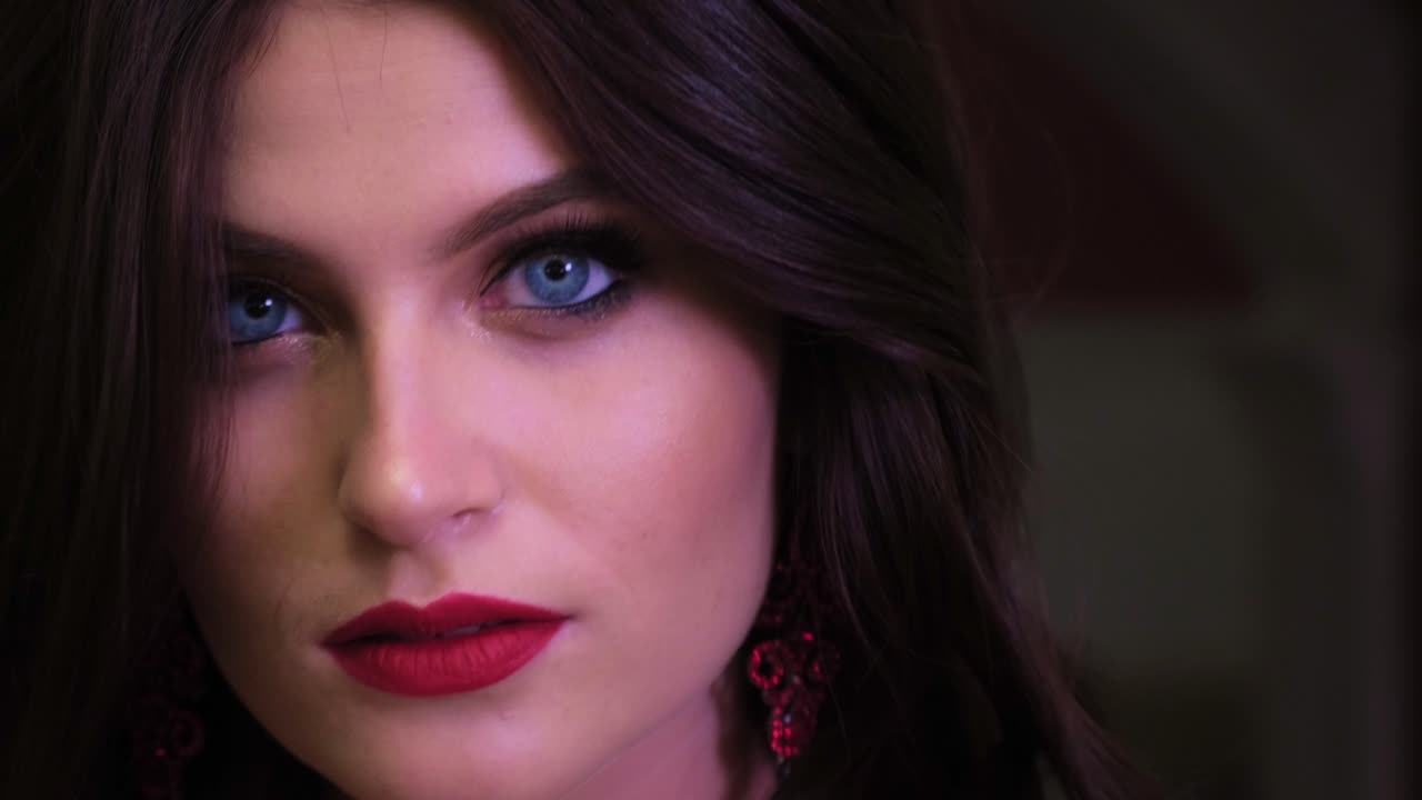 A Captivating Close-Up of a Woman with Striking Features and Bold Makeup, Highlighting Her Expressive Eyes and Vibrant Lip Color in a Dramatic Setting