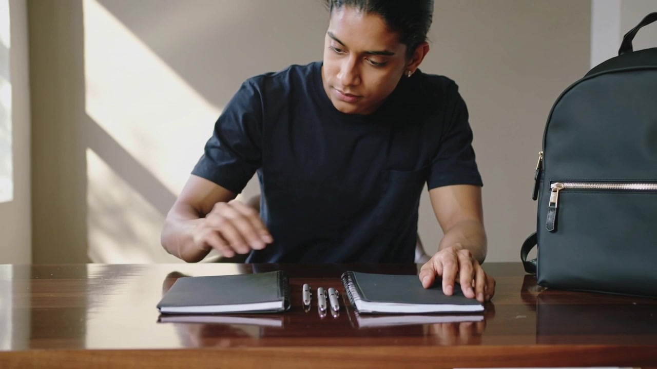 Leaning after resting hands, Asian teenager in black tee aligning pens, notebooks at table to study