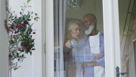 Worried-senior-diverse-couple-in-kitchen,-looking-through-window-and-embracing