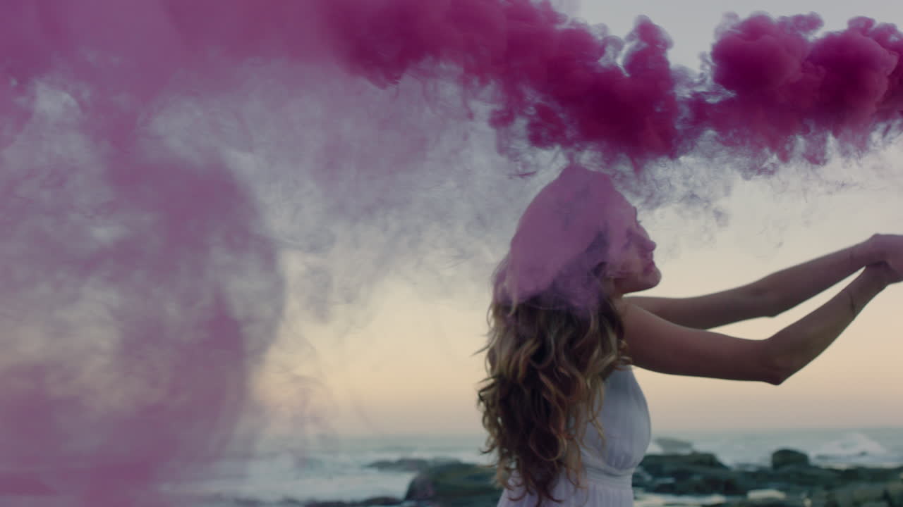 bellissima donna che agita una granata di fumo rosa che balla sulla spiaggia al mattino presto celebrando la libertà creativa