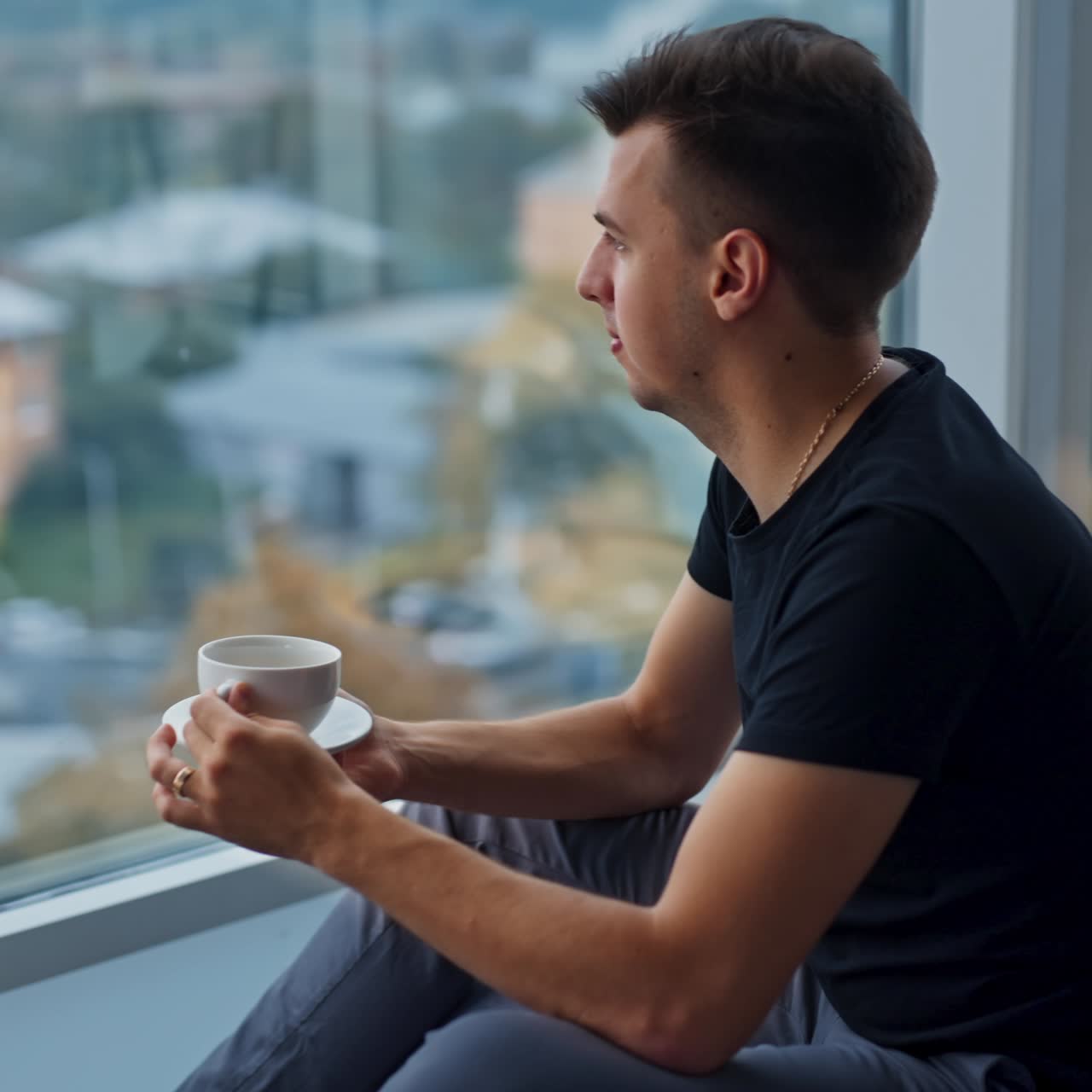 Brunet young man drinking coffee at lunch break sitting near panoramic window. Thoughtful male sitting in front of laptop. Blurred backdrop