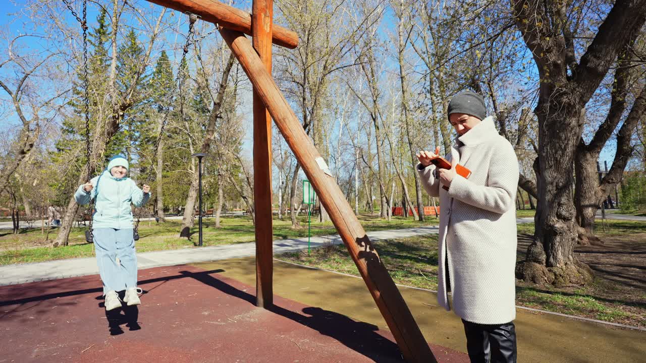 Giovane ragazza che dondola al parco mentre una donna usa il telefono