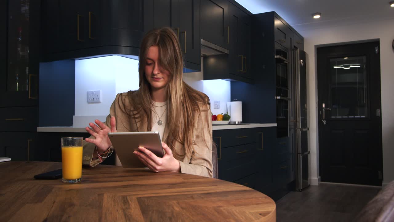 female model in a smart jacket drinking from a glass of orange juice using a iPad, tablet at a kitchen table in a modern kitchen