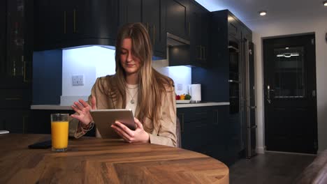 Modelo-Femenino-En-Una-Chaqueta-Inteligente-Bebiendo-De-Un-Vaso-De-Jugo-De-Naranja-Usando-Un-Ipad,-Tableta-En-Una-Mesa-De-Cocina-En-Una-Cocina-Moderna