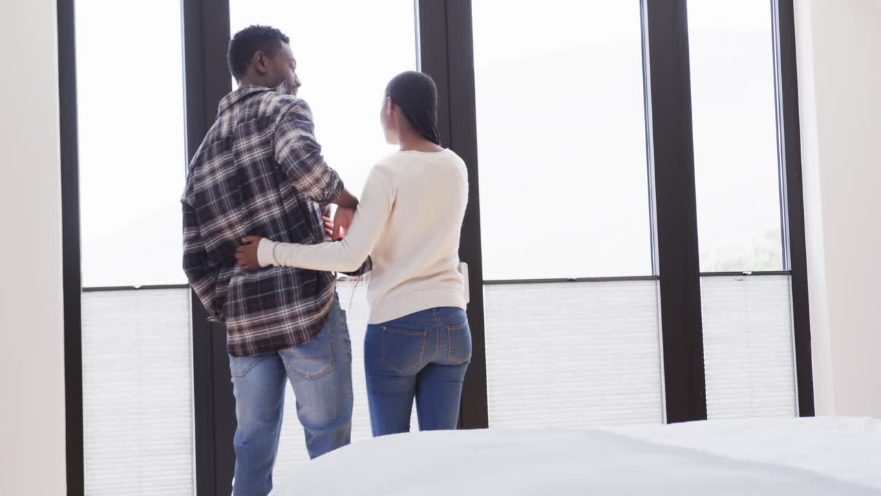 vista trasera de una feliz pareja afroamericana abrazándose frente a la ventana en casa, en cámara lenta