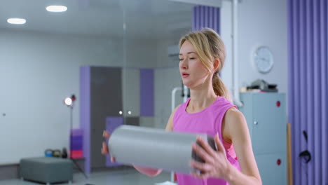 woman in pink workout attire lifts roller foam overhead, stretching arms upward in a well-equipped gym with musical device blinking light and various fitness equipment in the background