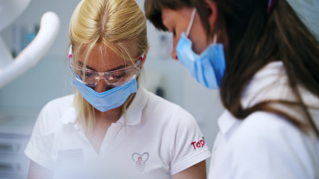 Blonde female medic wearing protective goggles and mask looks down at the patient. Dentist working in clinics.