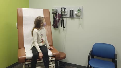 young girl sitting in the doctors office looking around curiously whilst waiting and wearing a face mask