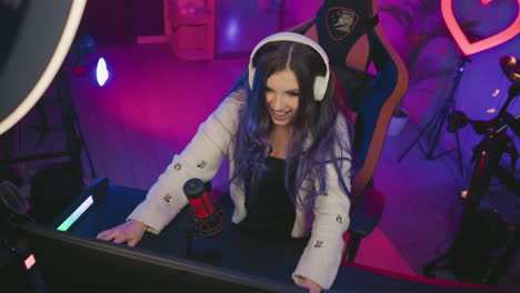 lady with long hair wearing headphones sits in gaming chair playing computer game, showing excitement after winning, surrounded by colorful lights, microphone, and music instruments in stylish studio