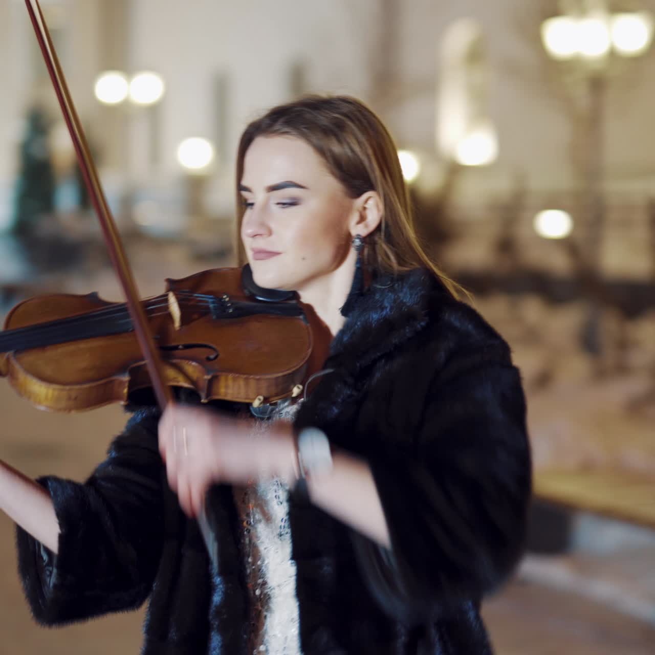 A young girl in a black coat is playing the violin on the background of buildings and evening lights in the centre of the city in the winter. Blurred Background