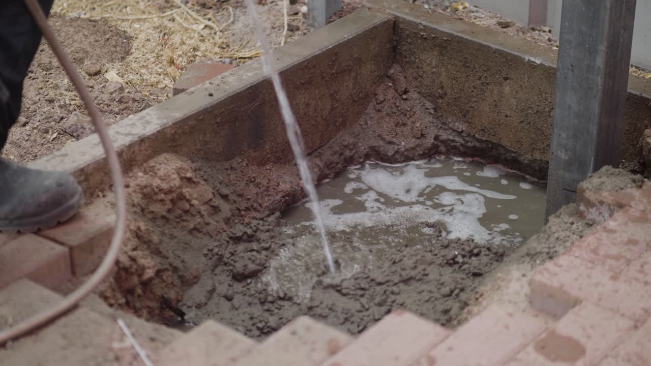 A construction worker adds water to cement in a hole, preparing the mixture for a building project. This footage captures the essential step of hydrating cement on a construction site.