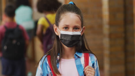 portrait of caucasian schoolgirl wearing face mask, standing in corridor looking at camera