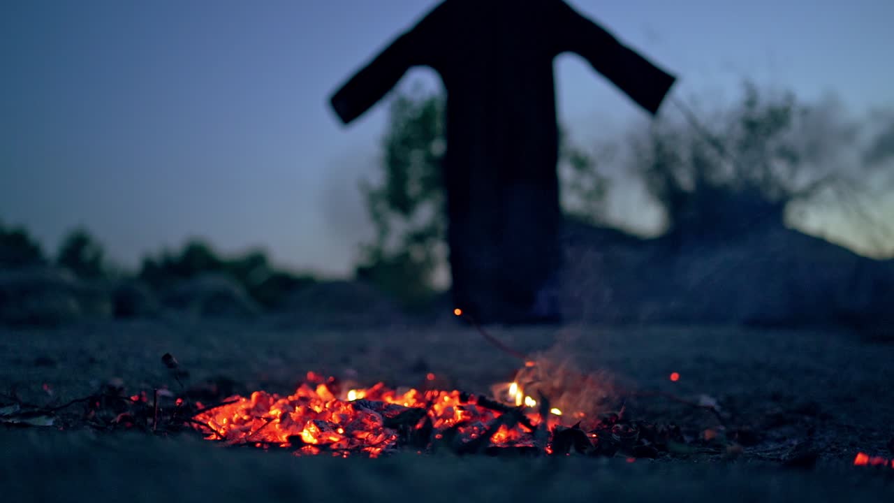 Embers of burnt-out fire and black witch outdoors. Dark death near smoldered logs in the evening. Mysterious figure on the natural background.